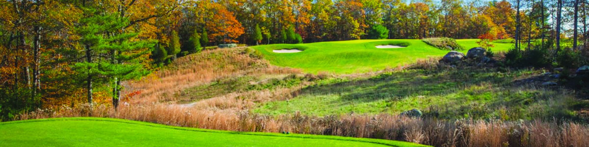 A scenic golf course with vibrant green grass, sand bunkers, rocks, and trees in fall colors under a blue sky with clouds.
