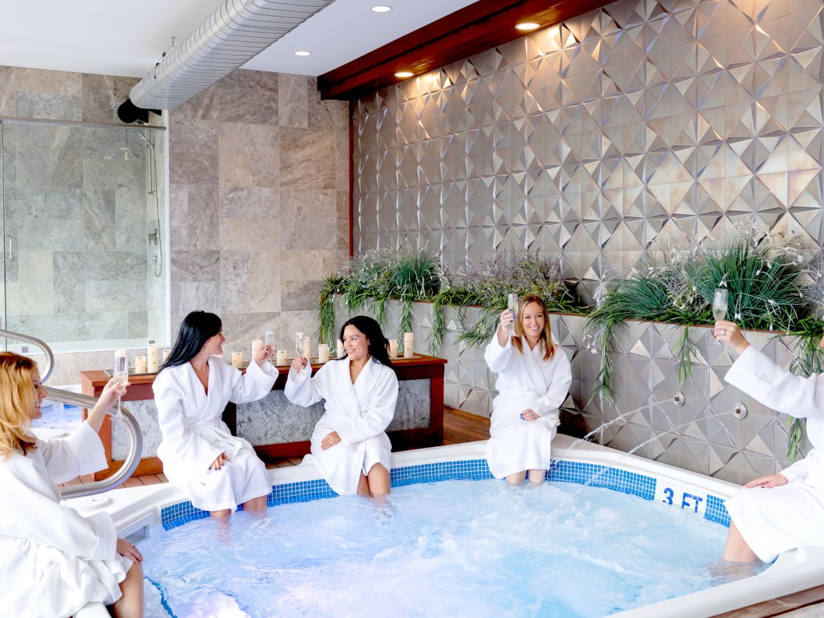 Five people in white robes sitting by a jacuzzi, holding drinks, and making a toast in a modern, indoor spa setting.