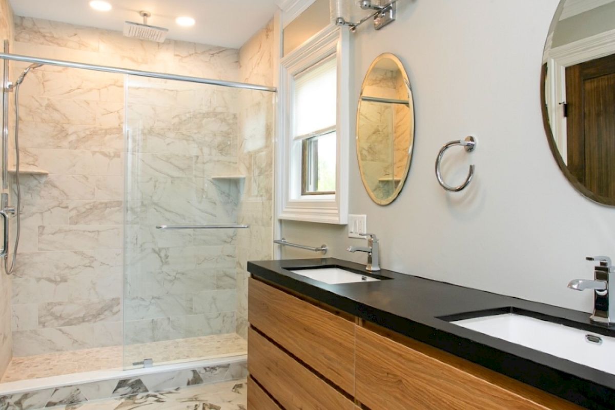 A modern bathroom with a glass shower, dual sinks, wooden vanity, and two round mirrors on the wall.
