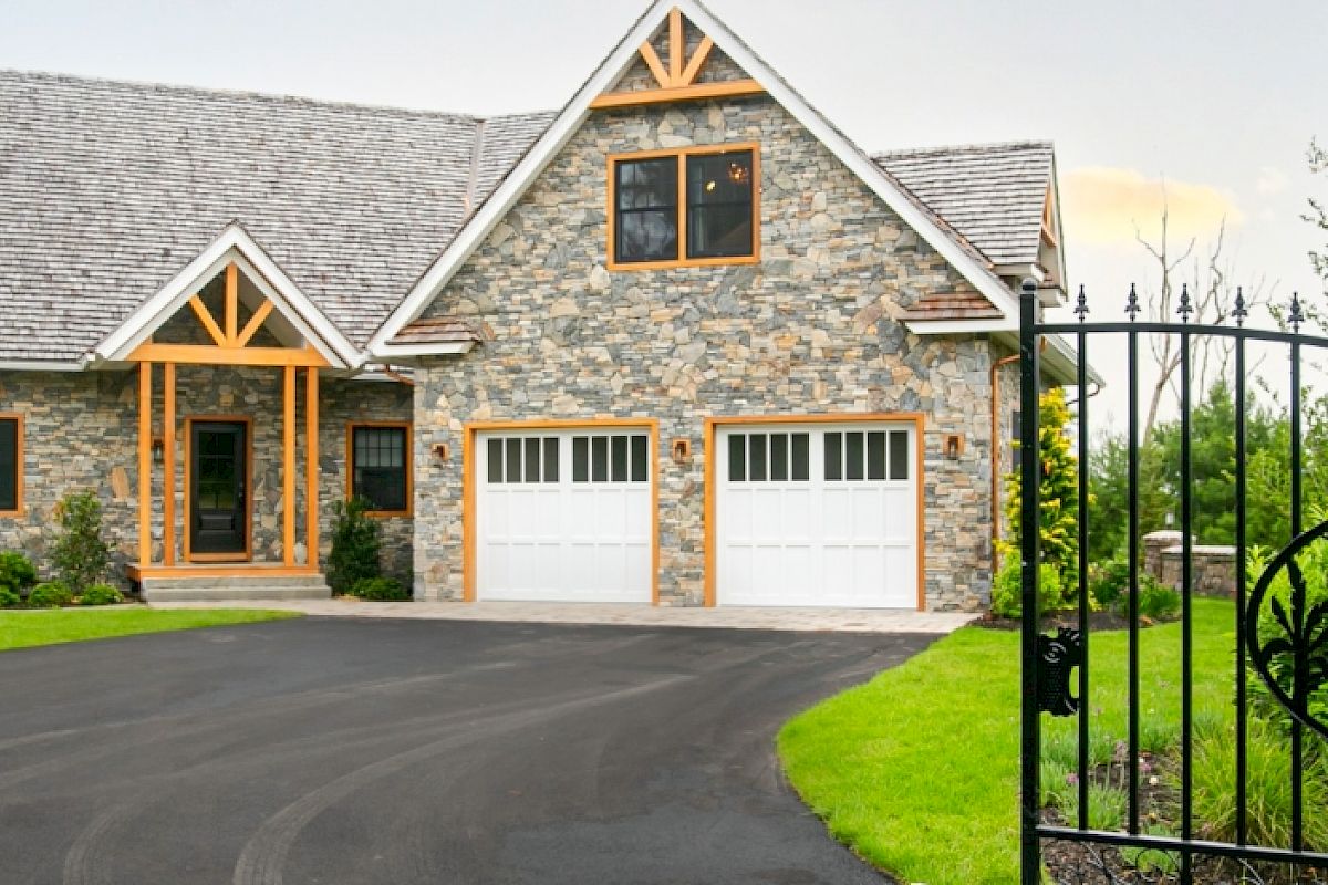 A stone house with a large driveway, two garage doors, and a decorative black gate featuring a deer design in a lush green setting.