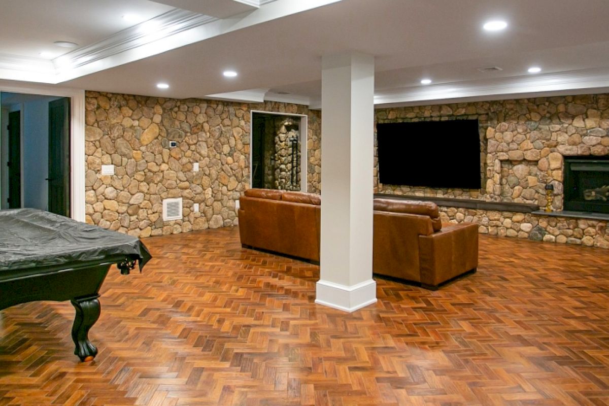 A basement with stone walls, featuring a pool table, brown couches, a TV, and a fireplace on a herringbone wood floor.