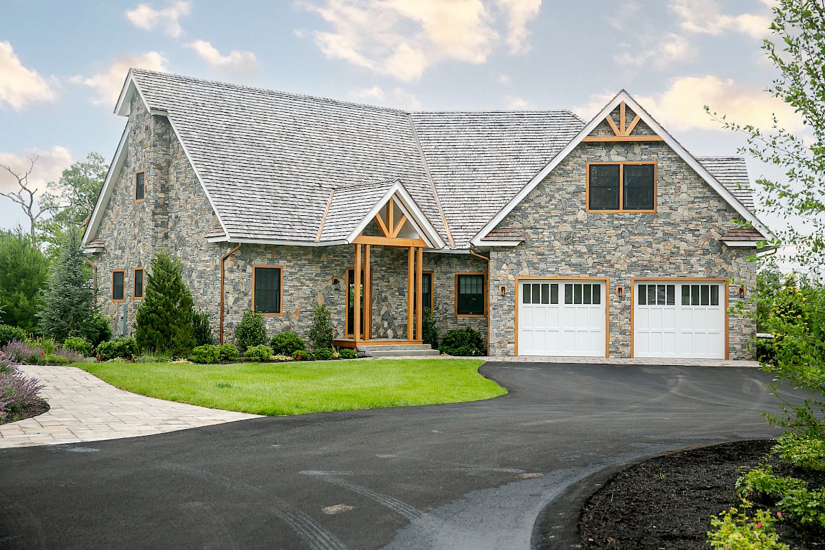 A modern stone house with a pitched roof, two garages, and a landscaped driveway surrounded by greenery and a clear sky.