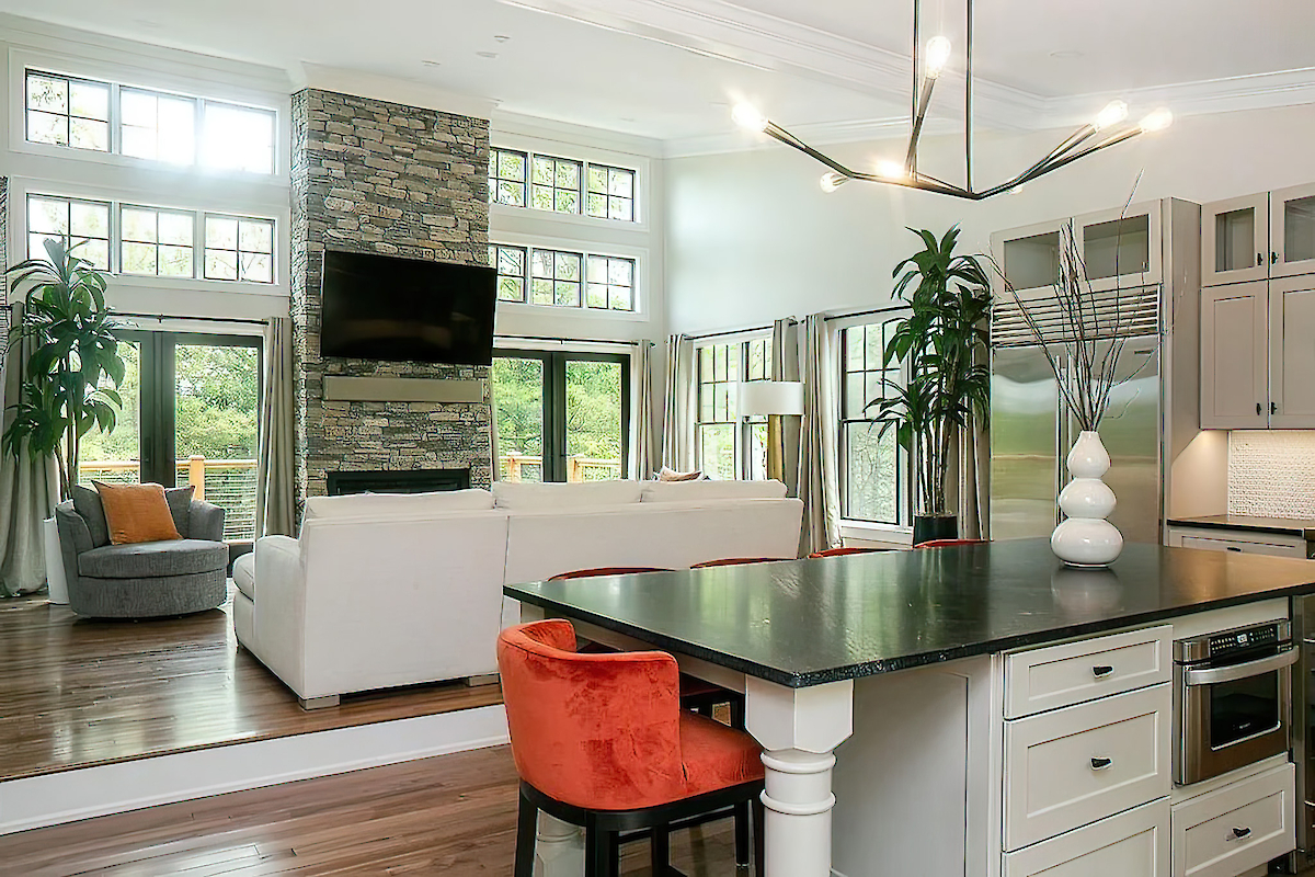 A modern living room and kitchen with large windows, stone fireplace, white and grey furniture, orange accents, and black countertop.