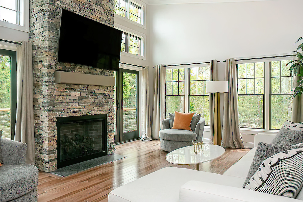 A modern living room with a stone fireplace, a mounted TV, large windows, a white sectional sofa, a round coffee table, and neutral-toned decor.