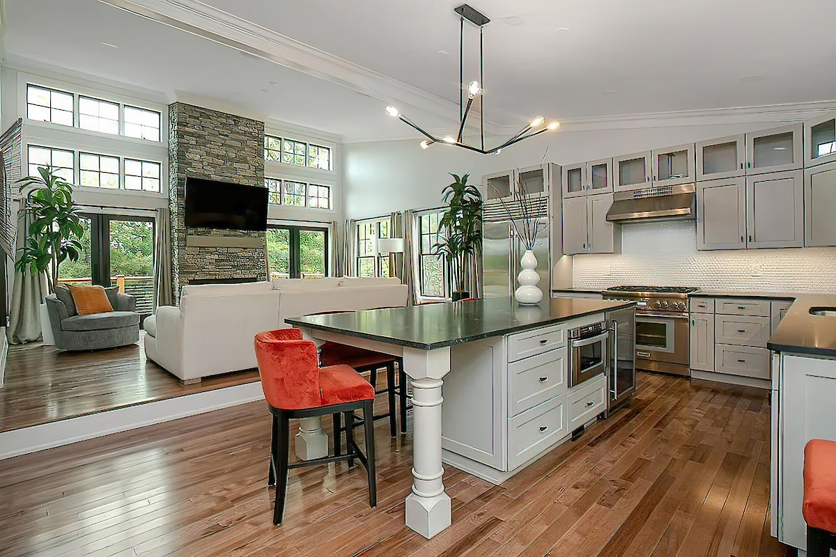 A modern kitchen with an island, bar stools, stainless steel appliances, and a connected living area with a fireplace and large windows.