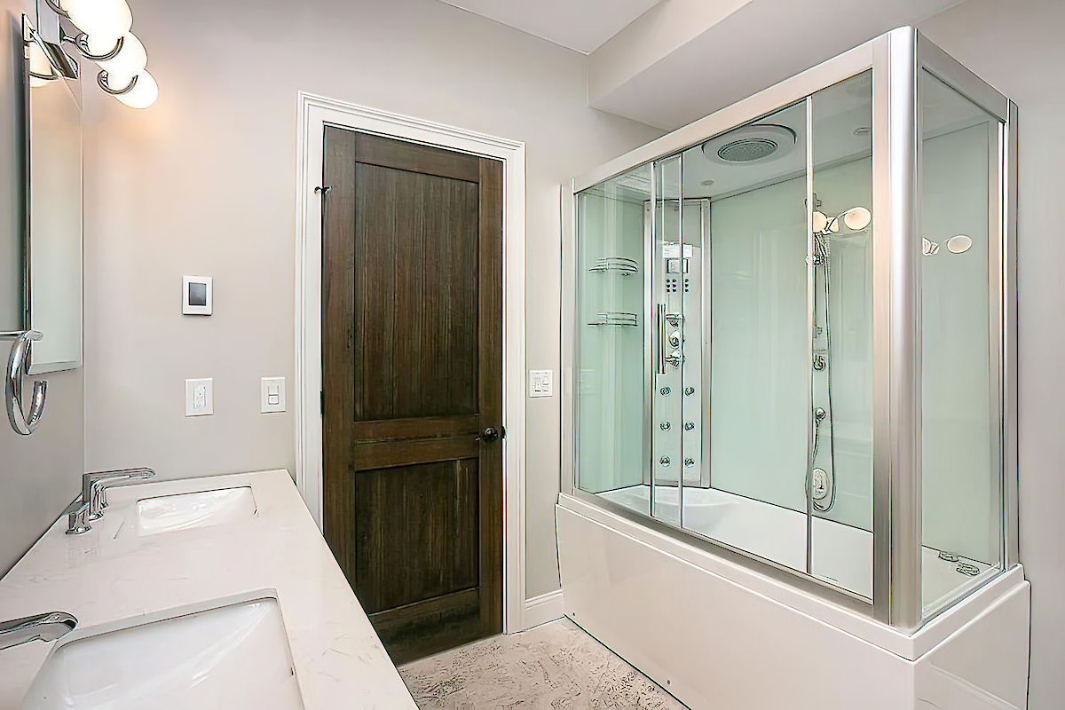 A modern bathroom with a double sink vanity, a dark wooden door, and a glass-enclosed shower/tub.