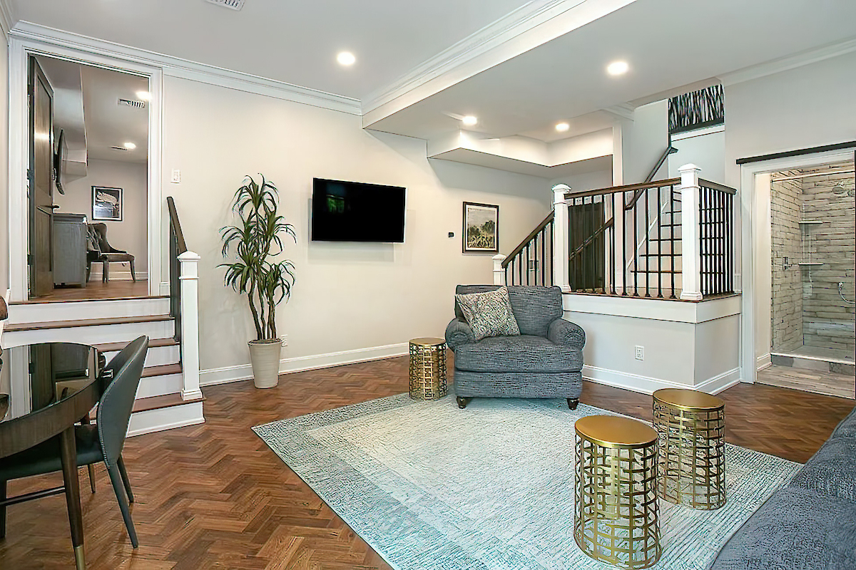 A modern living room with a grey armchair, TV, plants, and artwork. There are wooden stairs, a dining table, and three golden stools ending the sentence.