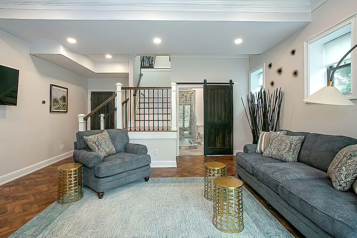 A modern living room with a gray couch, armchair, gold accent tables, and a TV. There's a staircase, a sliding door, and decorative wall pieces.