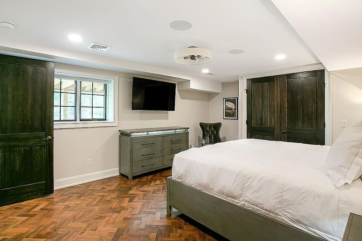 A bedroom with wood flooring features a bed, a dresser with a TV above it, a dark wood chair, and a closet with dark wooden doors.