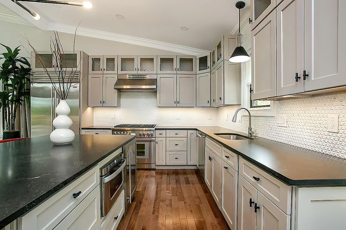 A modern kitchen with white cabinets, stainless steel appliances, a black countertop, wooden flooring, and various decor items in view.