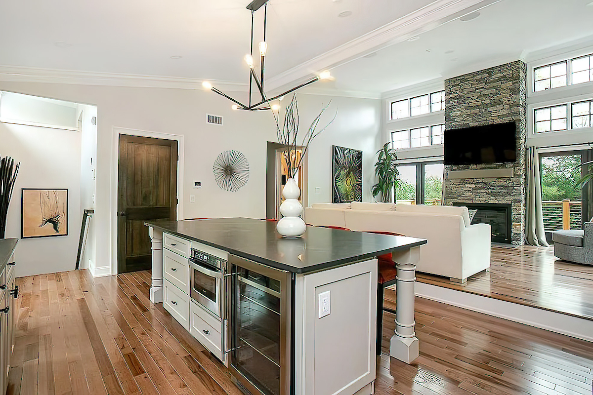 A modern open-concept kitchen and living area with a kitchen island, white sofa, stone fireplace, and large windows, featuring wooden floors.