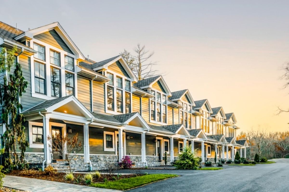 A row of modern townhouse units with peaked roofs, landscaped front yards, and a paved road winding in front, seen during early evening.