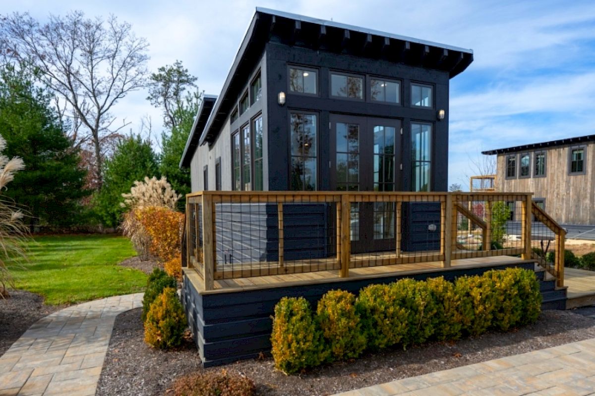 A modern tiny house with large windows and a wooden deck is set in a landscaped yard with greenery and a pathway.