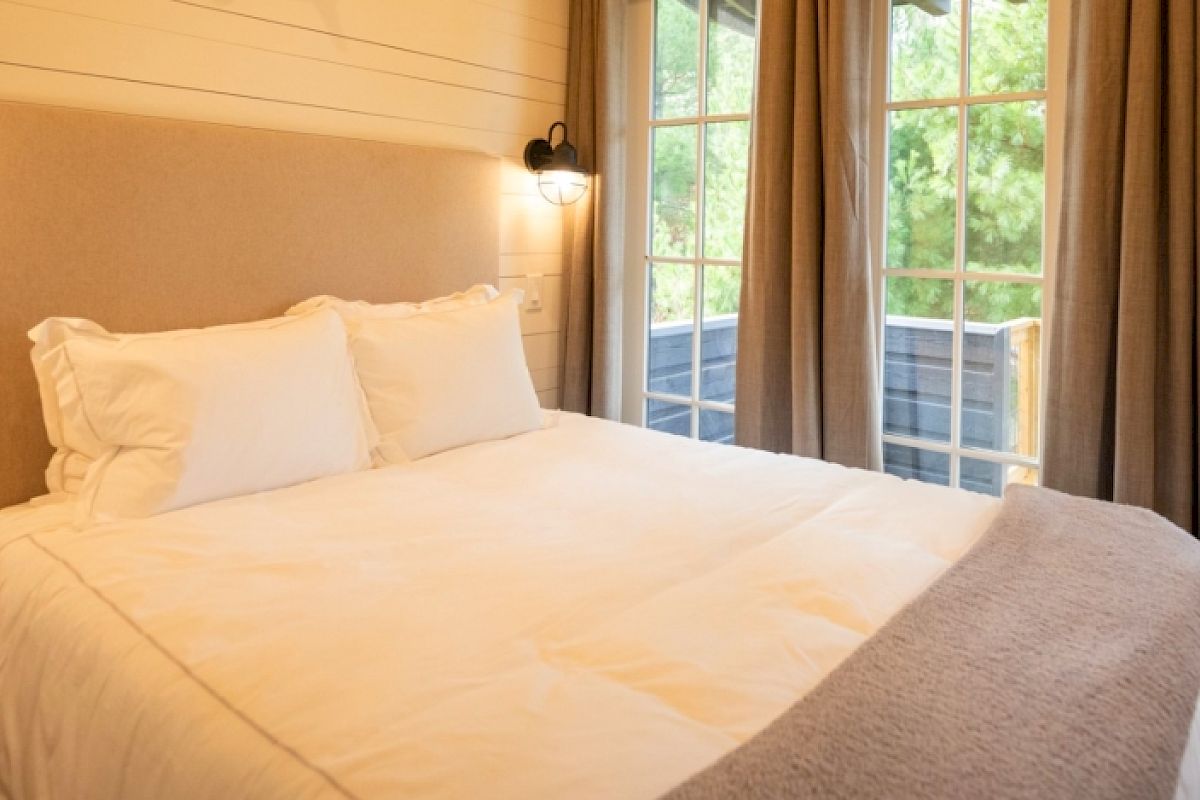 The image shows a cozy bedroom with a neatly made bed, soft lighting, and large windows with curtains offering a view of greenery outside.