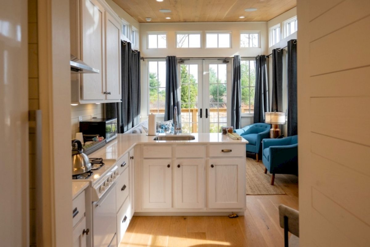 This image shows a compact, modern kitchen with white cabinets, leading to a cozy living area with blue chairs and large windows for natural light.