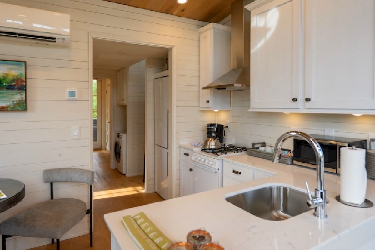 A modern small kitchen with white cabinets, sink, stovetop, and a dining table with chairs. Wall-mounted AC and art. Natural light enters the room.