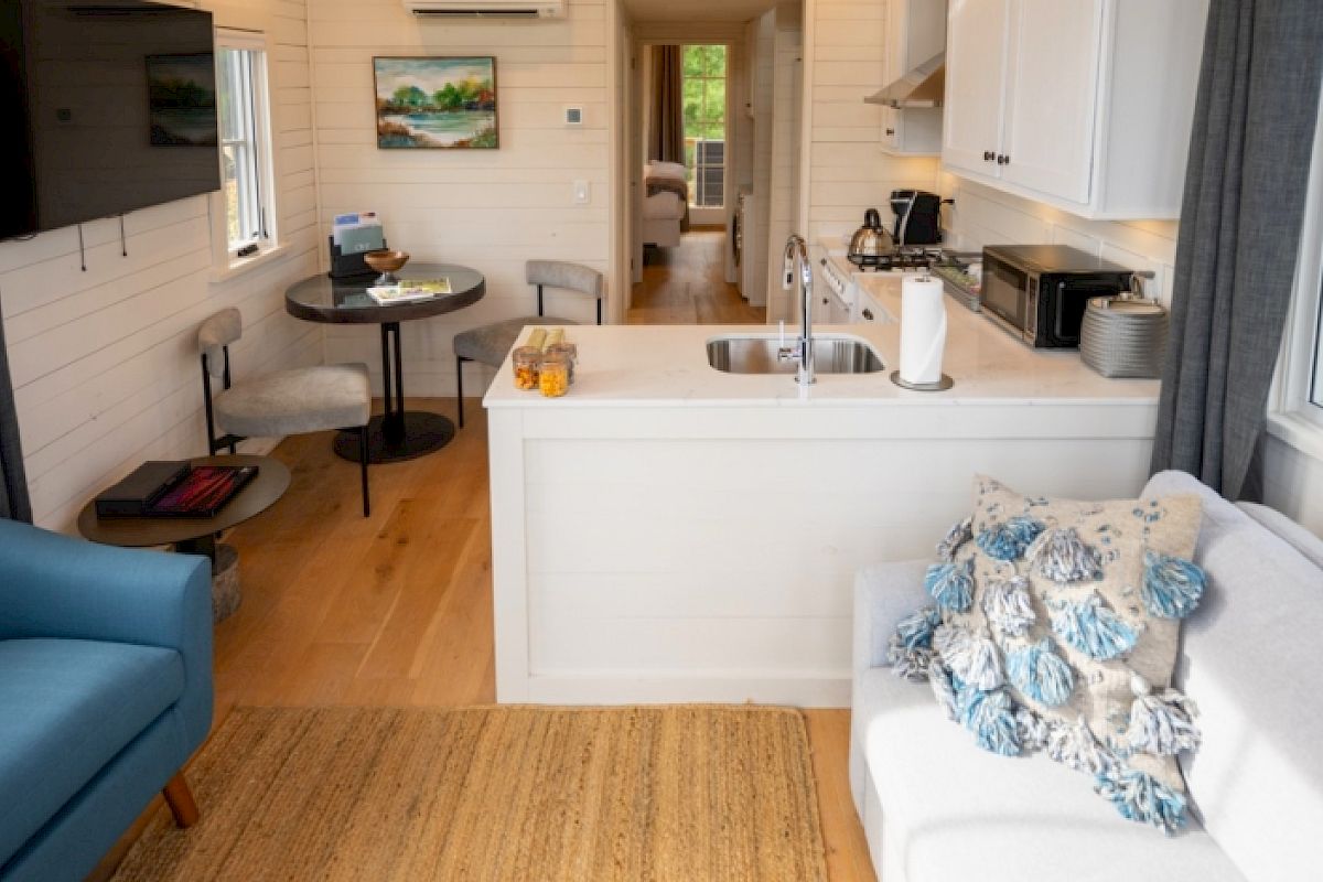 A cozy living space features a blue armchair, a white sofa with pillows, a small kitchen, a dining table, and a view of a bedroom in the back.