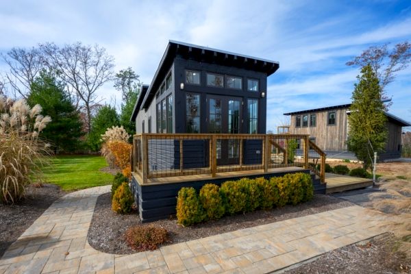 This image shows a modern tiny house with a wooden deck, surrounded by a stone pathway and landscaping.