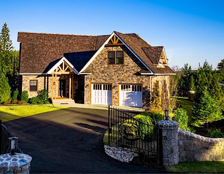 A stone house with a two-car garage, surrounded by trees and greenery, has a paved driveway and a gated entrance with stone pillars ending the sentence.