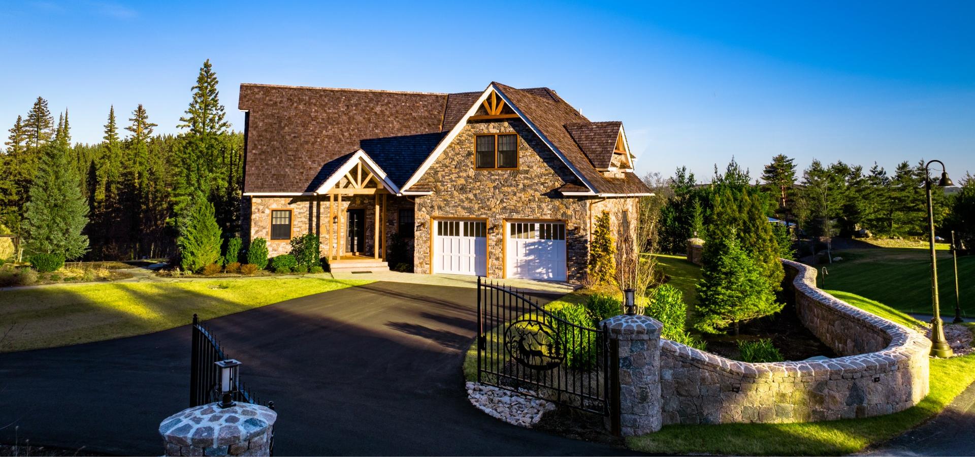 A stone house with a two-car garage, surrounded by trees and greenery, has a paved driveway and a gated entrance with stone pillars ending the sentence.