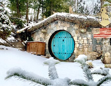 The image shows a snow-covered hobbit-like house with a stone facade, a circular blue door, a wooden owl sign, and trees surrounding the area.