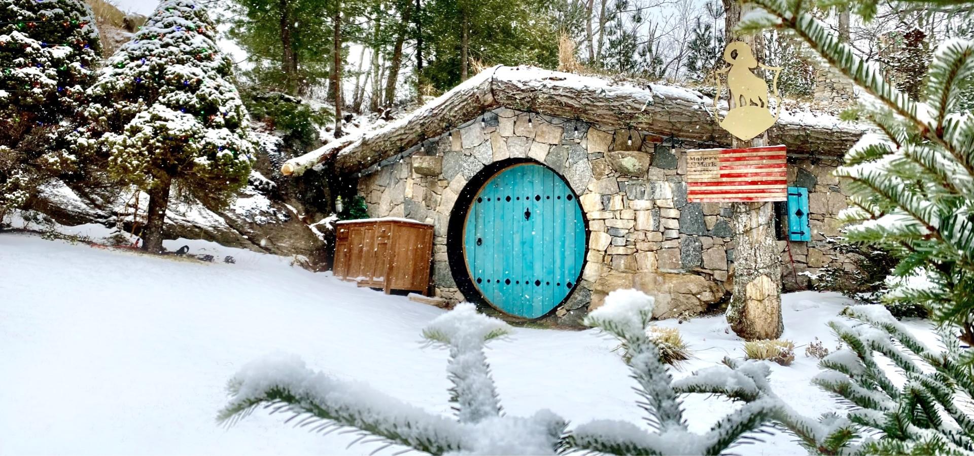 The image shows a snow-covered hobbit-like house with a stone facade, a circular blue door, a wooden owl sign, and trees surrounding the area.