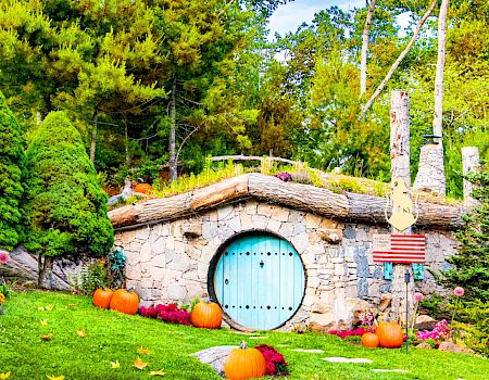 The image shows a small, charming, stone house with a round blue door, surrounded by greenery, trees, and pumpkins scattered on the lawn.