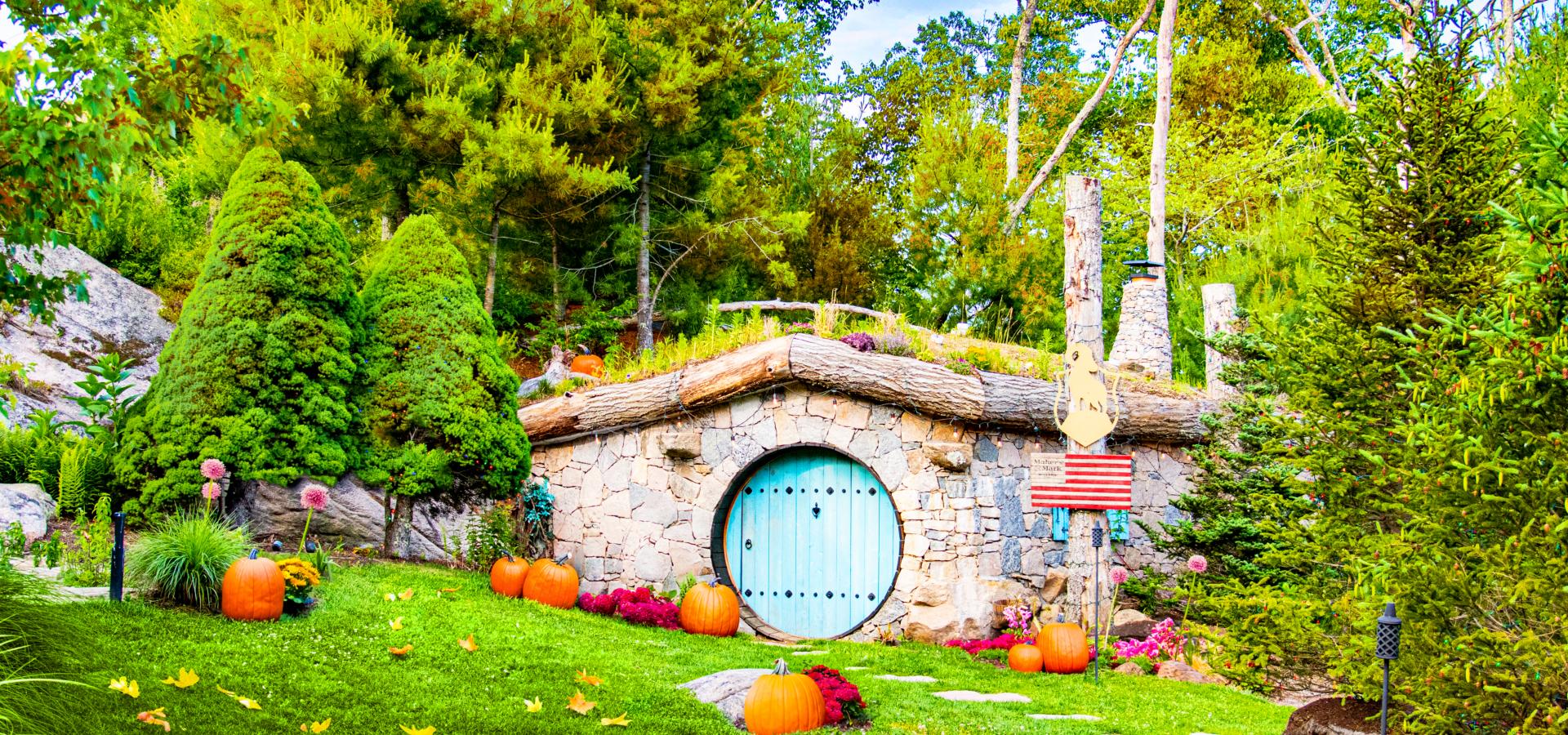 The image shows a small, charming, stone house with a round blue door, surrounded by greenery, trees, and pumpkins scattered on the lawn.