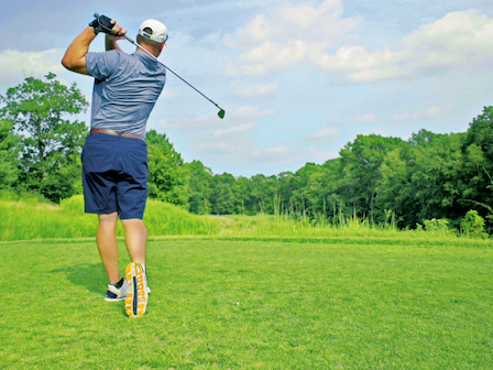 A golfer is shown from behind, swinging a club on a green course surrounded by trees under a partly cloudy sky.