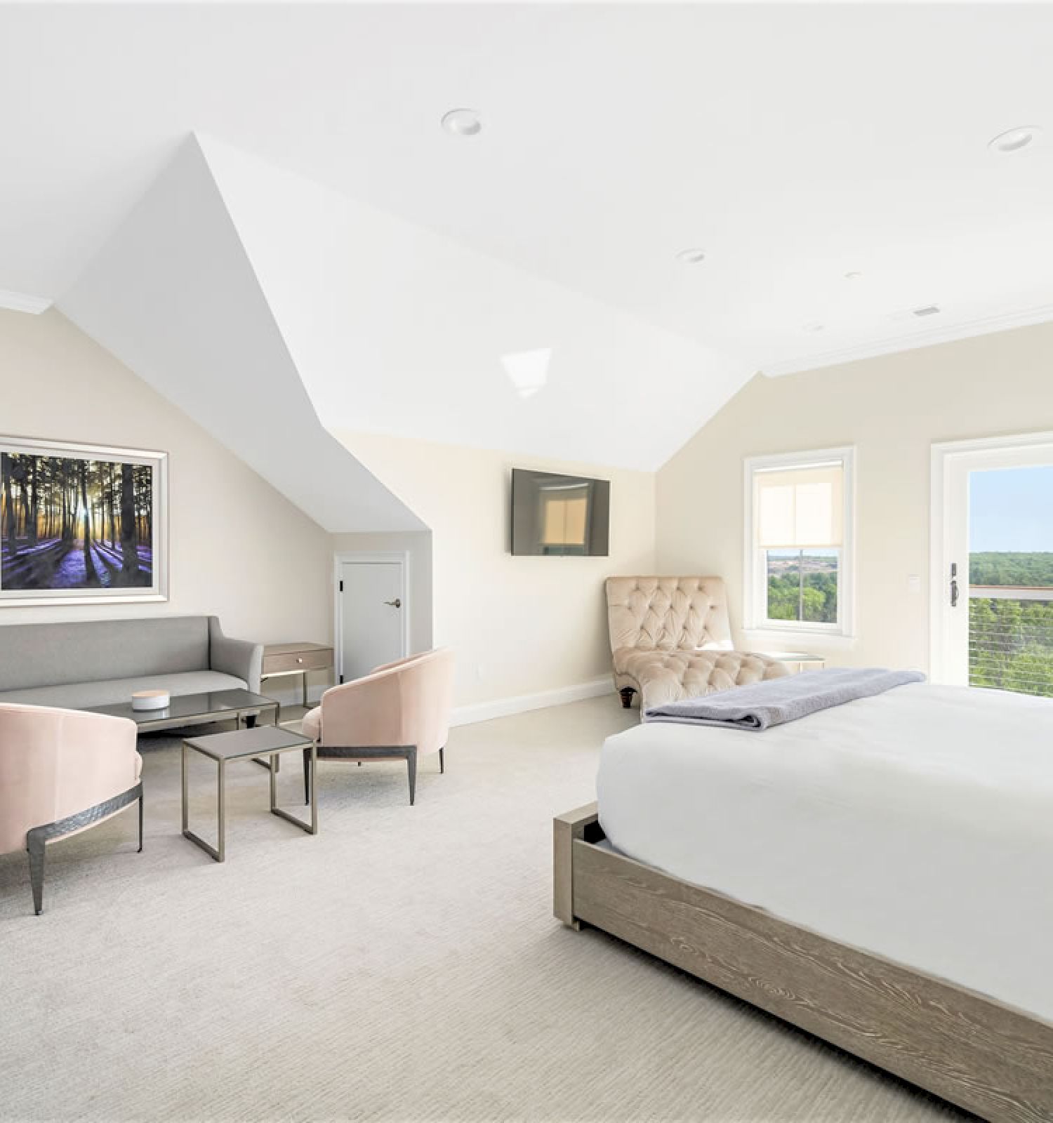A spacious, bright bedroom with a large bed, a sitting area with chairs and a sofa, wall art, a mounted TV, and a glass door leading to a balcony.
