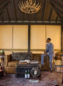 A man in a suit stands in a cozy, rustic room with antler chandelier, sofa, and coffee table. The decor includes globes and bottles on the carpet.