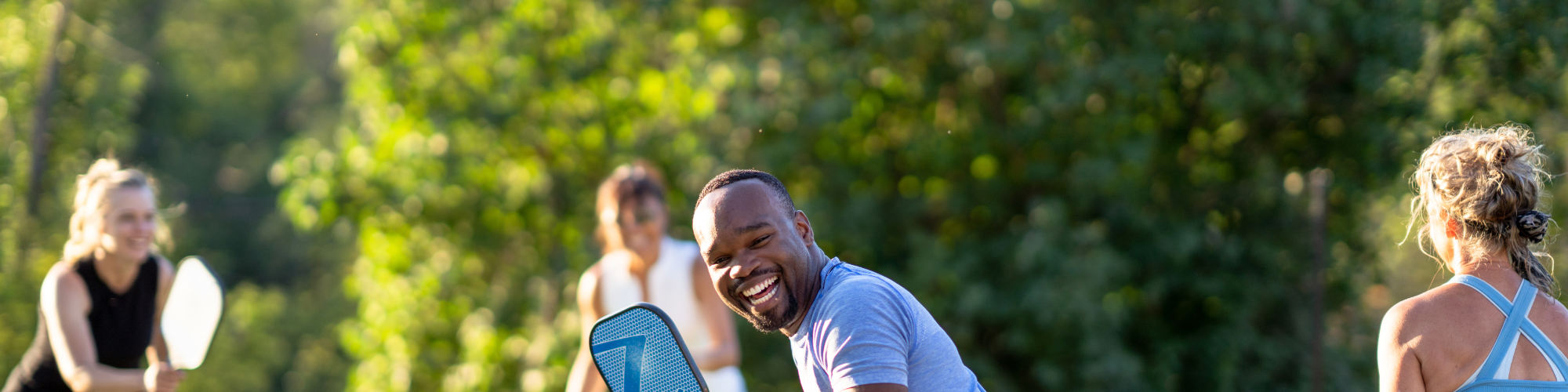 Four people are playing pickleball on an outdoor court, with green foliage in the background. One person is in action near the net, smiling.