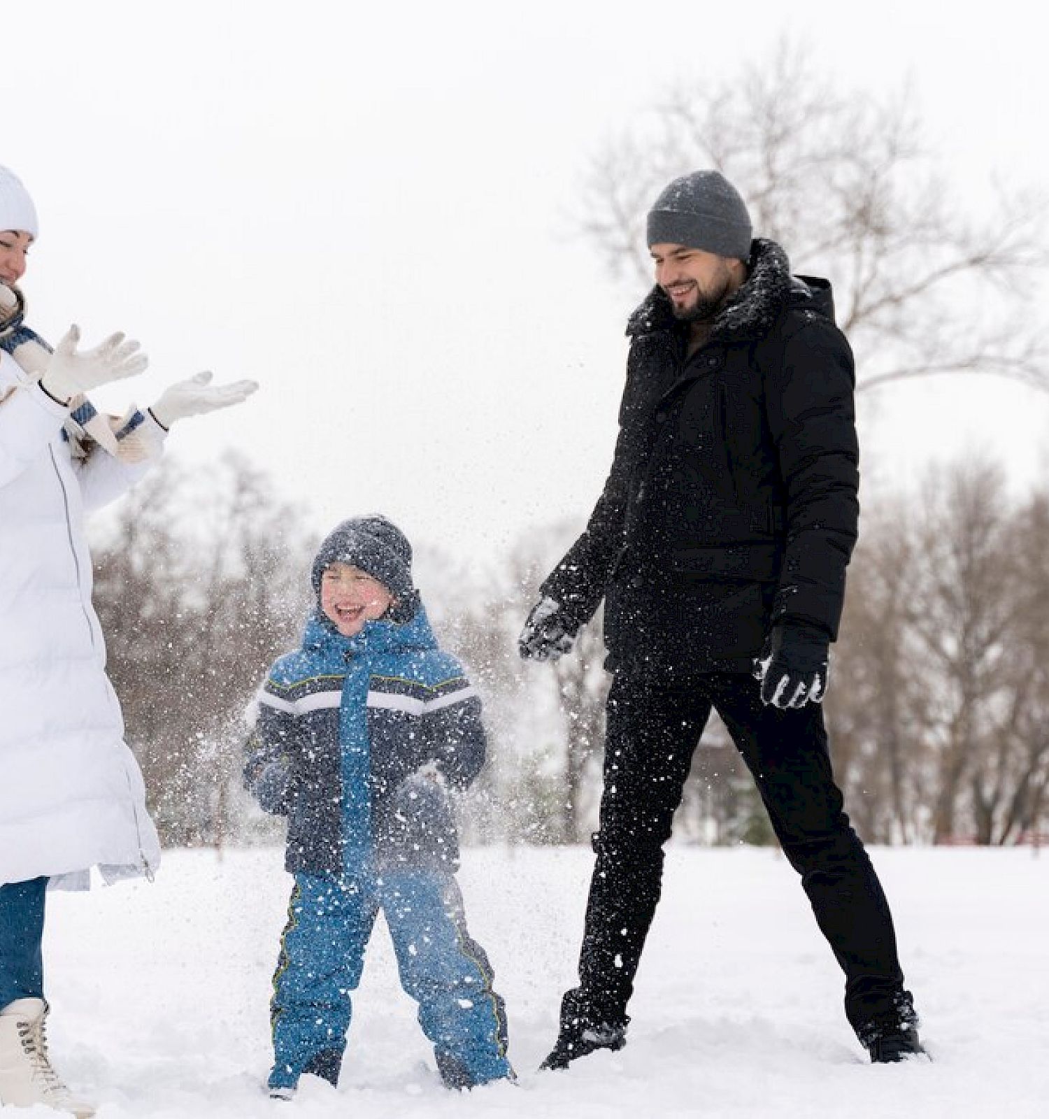 A family of three, dressed in winter clothing, enjoys playing in the snow outdoors, with trees in the background and snow falling around them.