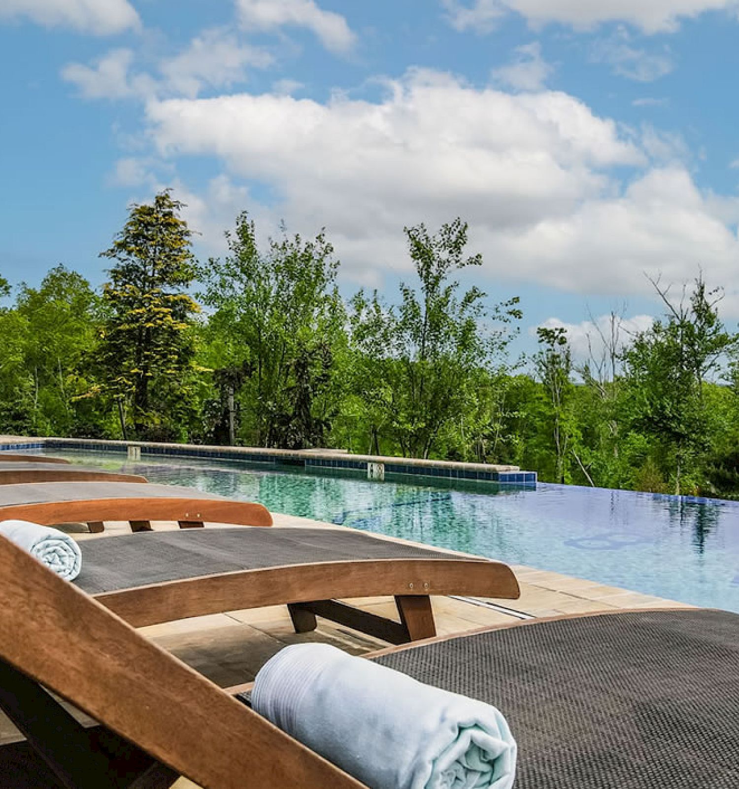 This image shows lounge chairs beside an outdoor infinity pool surrounded by trees and a blue sky with some clouds.