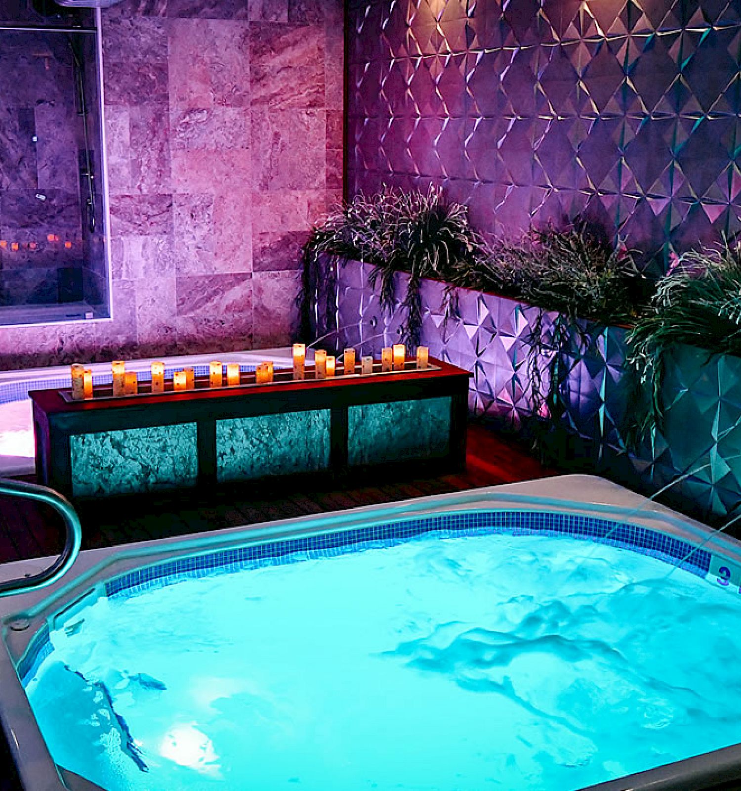 The image shows a luxurious indoor spa area with two jacuzzis, candles, decorative plants, and contemporary wall designs.