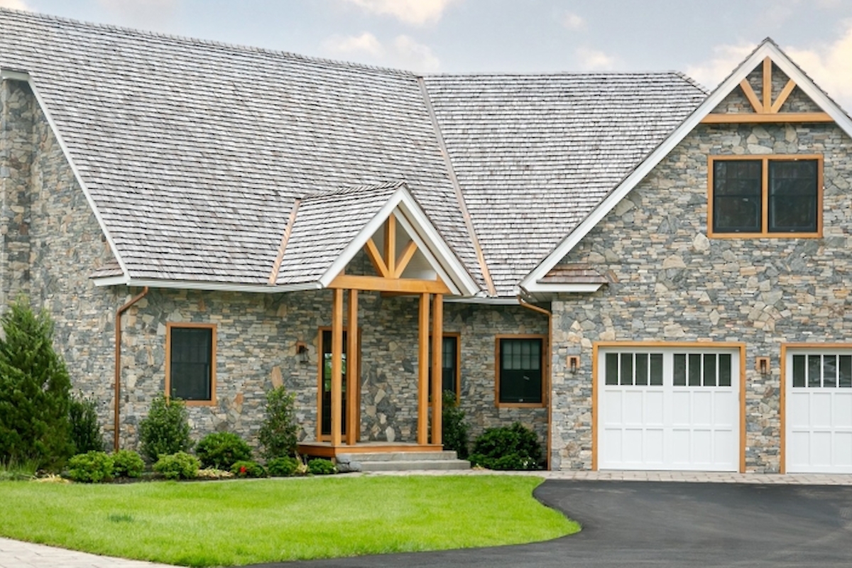 A large stone house with a gabled roof, a wooden front entrance, manicured lawns, and a dark asphalt driveway leading to double garage doors.