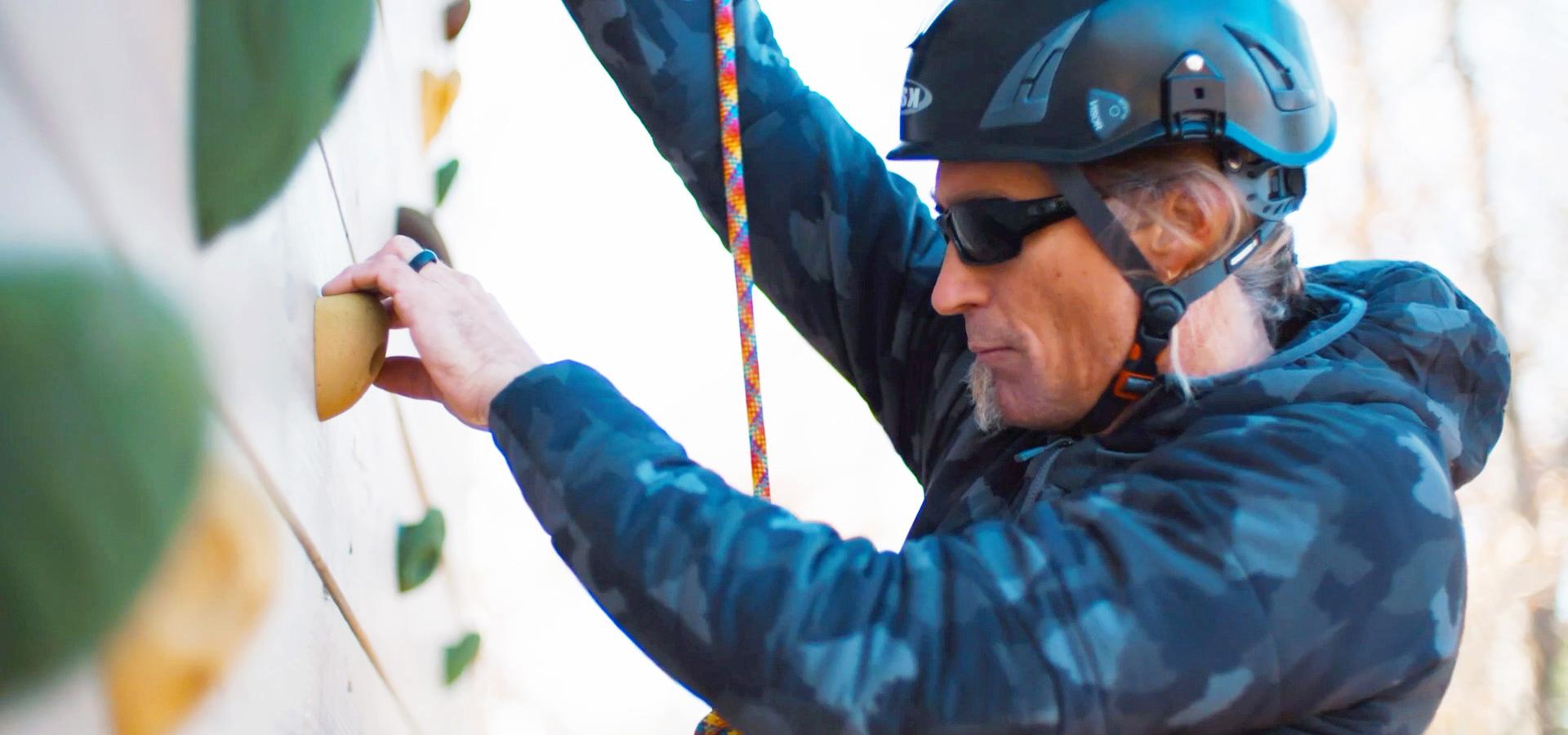A person wearing a helmet and sunglasses climbs a wall with handholds, using a rope for safety. They appear focused on the activity.