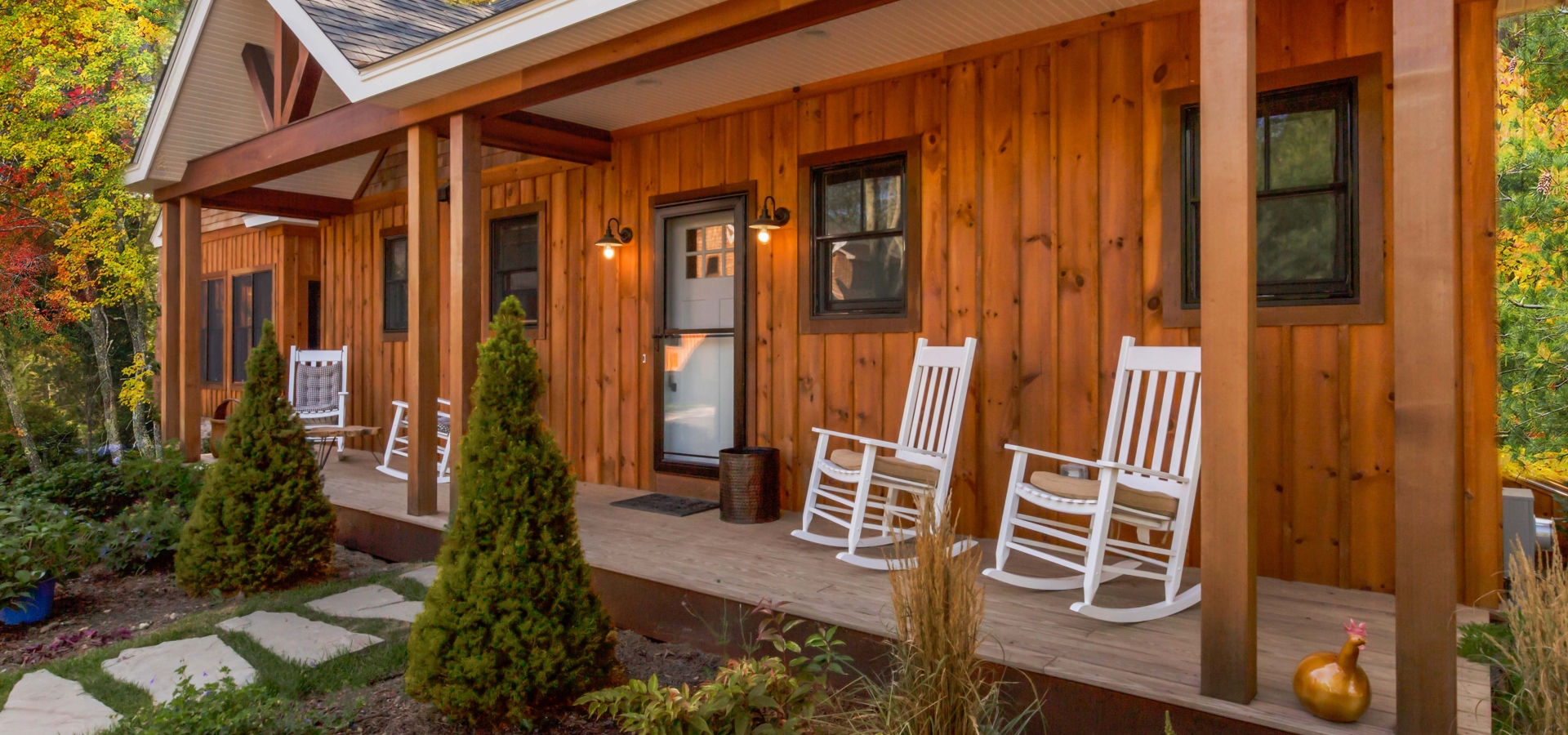 A wooden house with a porch, white rocking chairs, small trees, and a jug on the porch corner.