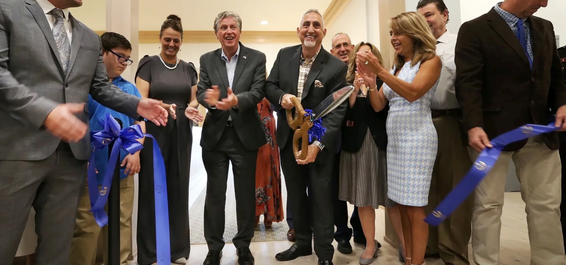 A group of people stand together at a ribbon-cutting ceremony, with one person holding large scissors and others clapping.