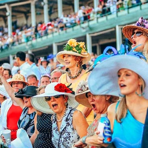 A group of women wearing large, colorful hats cheer and watch an event at a crowded outdoor stadium.