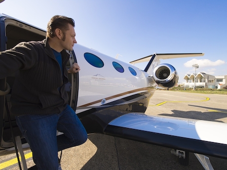 A person is stepping out of a private jet on the tarmac, with the airport terminal visible in the background and a clear sky overhead.