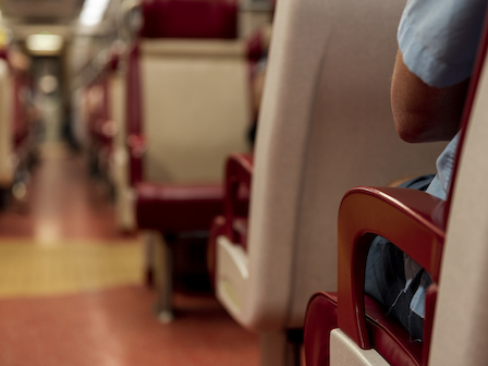 The image shows the interior of a train with red and beige seats. A person in a blue shirt is seen from behind, with their arm resting on the seat.
