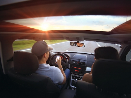 Two people are in a car with a sunroof, driving during sunset; the driver is using a phone while driving on a highway.