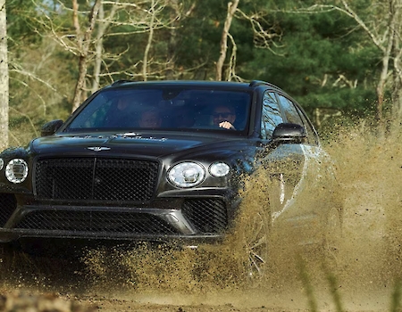 A black SUV is driving through a muddy, off-road environment with mud splashing up around it, in a wooded area.