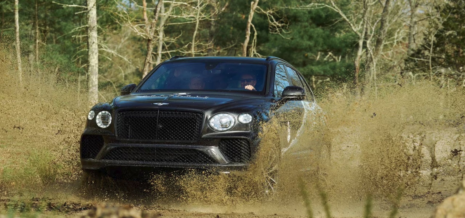 A black SUV is driving through a muddy, off-road environment with mud splashing up around it, in a wooded area.
