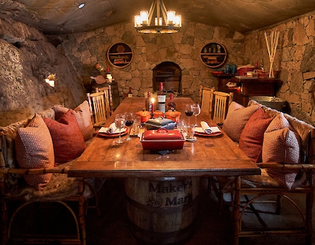 This image shows a cozy dining room with stone walls, a wooden table surrounded by chairs with cushions, shelves, and a chandelier overhead.