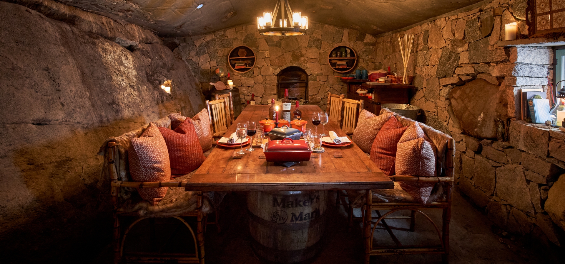This image shows a cozy dining room with stone walls, a wooden table surrounded by chairs with cushions, shelves, and a chandelier overhead.