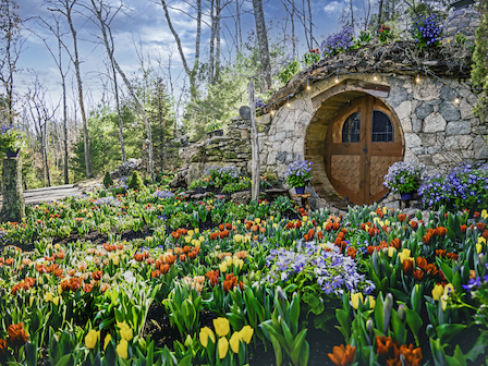 A small stone house with a round wooden door is surrounded by colorful flowers and trees, resembling a hobbit home.