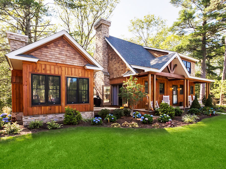 A charming wooden house with a brick chimney surrounded by greenery and vibrant flowers under a clear, sunny sky.