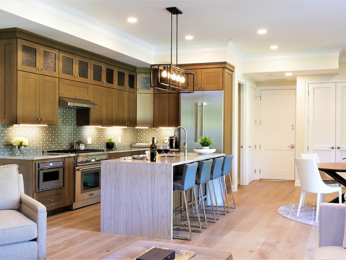 A modern kitchen with an island, bar stools, stainless steel appliances, and wooden cabinets, adjacent to a dining and living area.